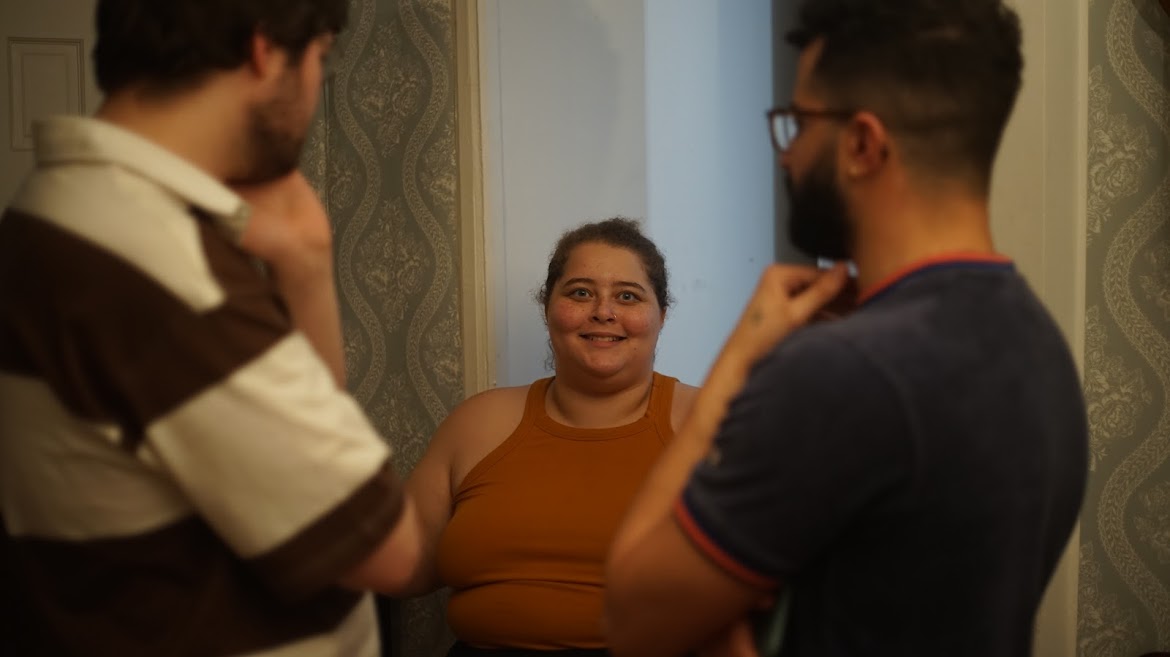 Amelie wears an orange
            tank top and stands center frame, smiling at the viewer. Her collaborators 
            Jon and Kevin stand blurred in the foreground, looking straight at her.